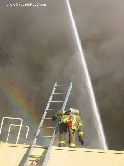 A rainbow of hope (bottom left) brings a bit of hope as a fireman looks on, while a ladder truck forcefully shoots tons of water down onto the burning building.